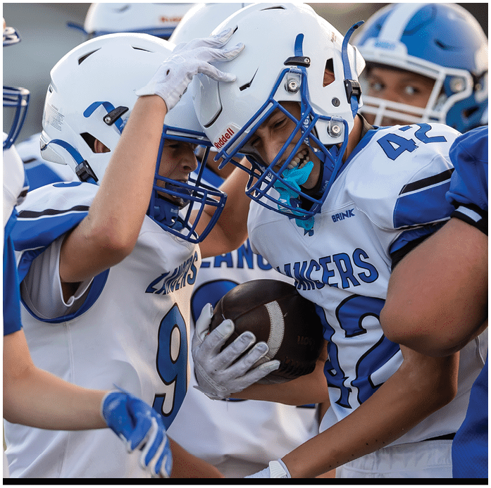 Two people wearing white football helmets and jerseys are celebrating with their foreheads touching and smiling. Description generated by AI