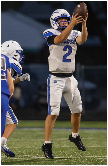 A person wearing a white football helmet and a white jersey with blue accents is reaching out to catch a football. Description generated by AI