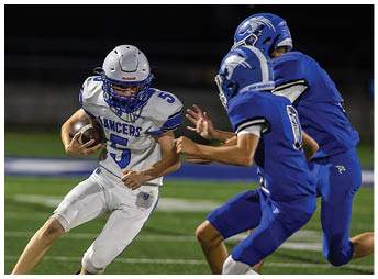 Two people in blue football helmets and one person in a white football helmet are engaged in a play on a football field at night. Description generated by AI