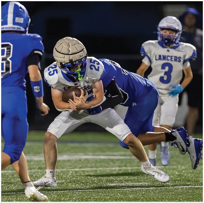 Two people in football uniforms are engaged in a tackle during a nighttime game with one holding the ball and another player in the background watching. Description generated by AI