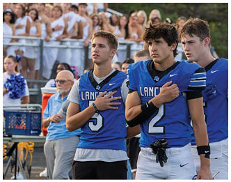 Adults and teenagers in blue football uniforms stand on a field with their hands over their hearts while a crowd of people dressed in white watches from the bleachers. Description generated by AI