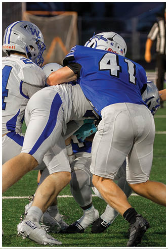 Four people in football uniforms are engaged in a tackle on a green field with a referee in the background. Description generated by AI