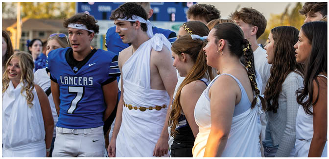 A group of people dressed in a mix of sports uniforms and ancient Greek-style togas are gathered together outdoors near a scoreboard. Description generated by AI