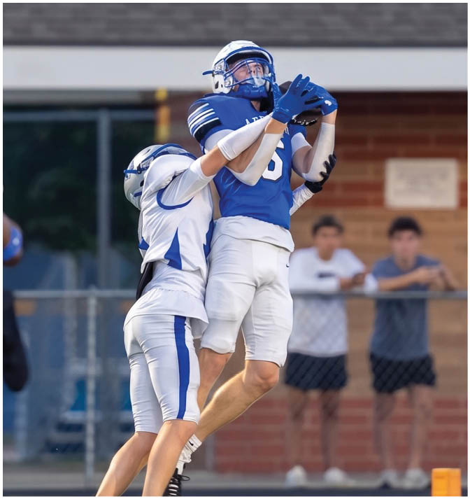A person in blue football gear is catching a ball while two people in white football uniforms are nearby on a field with a building and fence in the background. Description generated by AI