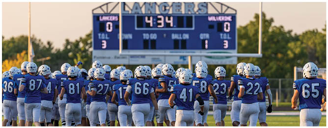 A group of people wearing blue football uniforms and helmets stand on a field facing a scoreboard showing a tied score. Description generated by AI