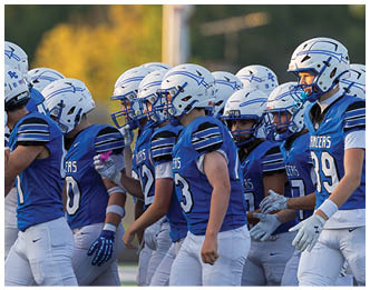 A group of people wearing blue sports jerseys, white helmets, and white pants are walking together on a green football field. Description generated by AI