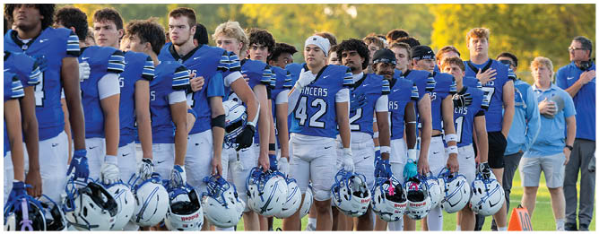 A group of people wearing blue and white football uniforms stand in a line outdoors with helmets on the ground in front of them and coaches standing nearby. Description generated by AI