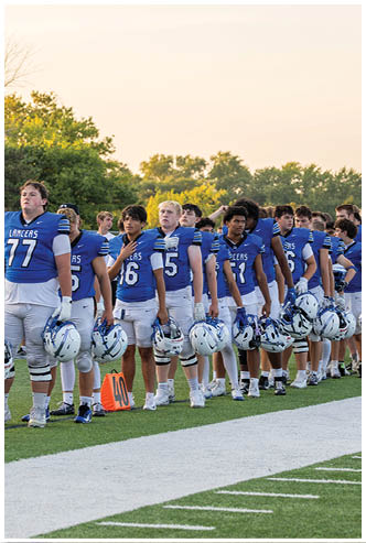 A group of people wearing blue football uniforms stand in a line on a football field holding their helmets, with some placing their hands over their hearts. Description generated by AI