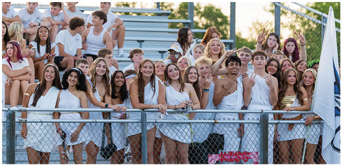 A group of people dressed mostly in white are gathered closely together behind a chain-link fence, smiling and posing for a photo in an outdoor setting with bleachers and trees in the background. Description generated by AI