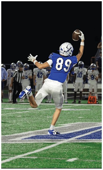 A person wearing a blue football jersey with the number 89 is jumping and reaching up to catch a football on a field with other players in the background. Description generated by AI
