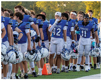 A line of football players in blue jerseys and white pants stand on the sideline of a football field holding their helmets during what appears to be a pre-game or halftime moment. Description generated by AI