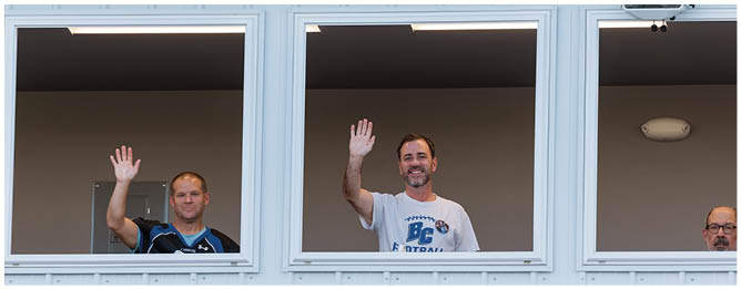 Three adults are standing behind a white railing with three open windows, two of them waving and one looking forward. Description generated by AI