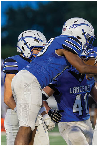 People wearing blue and white football uniforms are engaged in a game, with one player holding the ball and others preparing to block or tackle. Description generated by AI