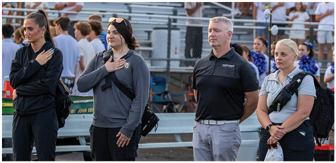 Adults and young people stand on a sports field with their hands over their hearts while a crowd watches from bleachers in the background. Description generated by AI