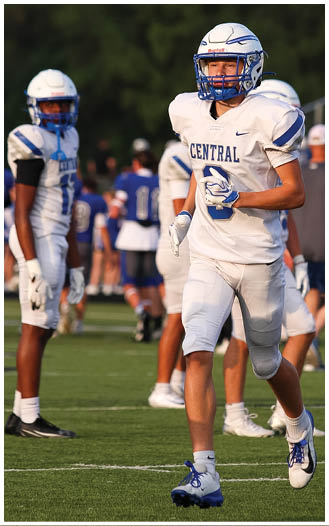 A person wearing a white football uniform with “CENTRAL" on the jersey is standing on a field with other players and people in the background. Description generated by AI