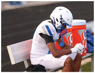 A person wearing a white football helmet and uniform is sitting on a bench next to orange water coolers on a sports field. Description generated by AI
