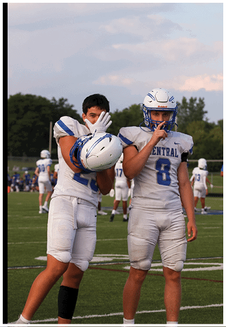 Two people in football uniforms stand outdoors with one holding a helmet and the other making a shushing gesture. Description generated by AI