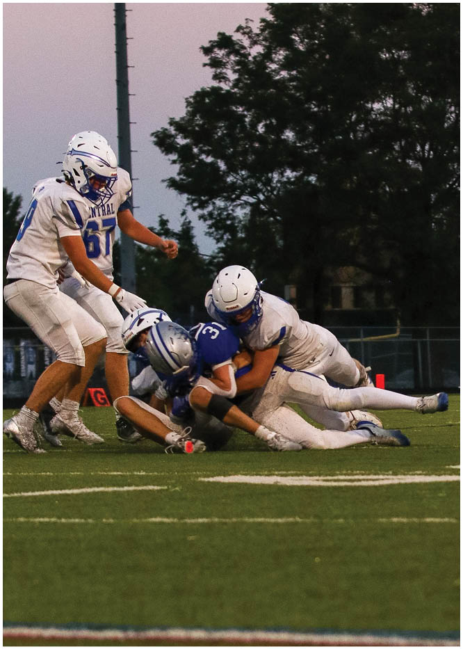 Three people in football uniforms are on a field with two players tackling a third player wearing a blue jersey. Description generated by AI