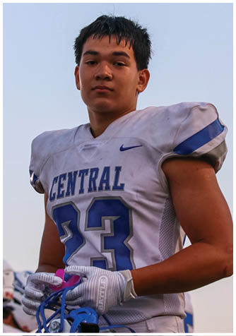 A person wearing a white sports jersey with blue accents is looking toward the camera against a clear sky background. Description generated by AI
