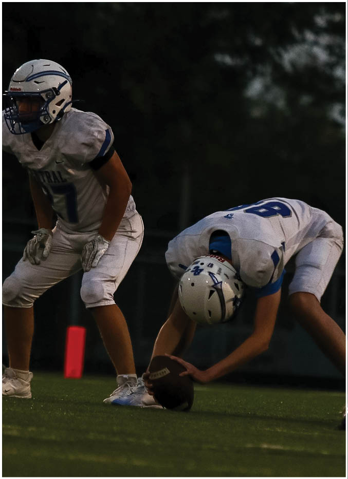 Two people in football uniforms and helmets are positioned on a field, with one holding a football ready to snap it and the other standing in a ready stance. Description generated by AI