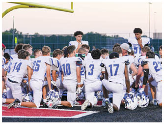 A group of people wearing white football uniforms with blue numbers are kneeling on a field under a goalpost, appearing to listen to someone standing among them. Description generated by AI