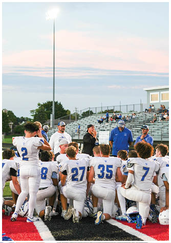 A group of people in white sports uniforms kneel and stand on a football field near the sideline while coaches in casual clothing talk to them during a game or practice at dusk. Description generated by AI