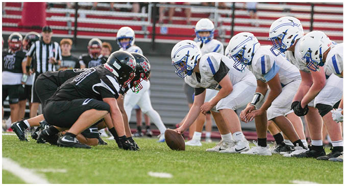 Two opposing American football teams in black and white uniforms are lined up at the line of scrimmage on a grassy field with players crouched and ready for the play to start. Description generated by AI