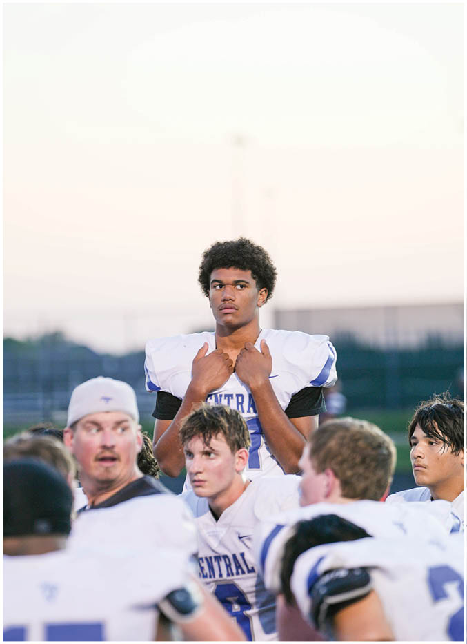 A group of people in football uniforms are gathered together outdoors with one person standing behind the others looking focused. Description generated by AI