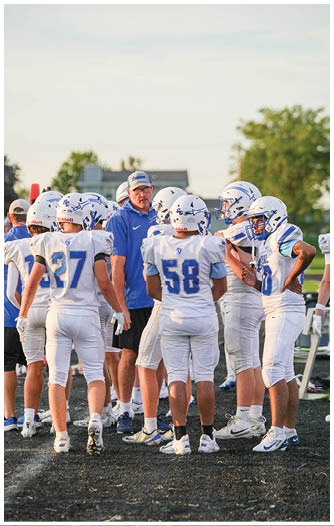 A group of people wearing white football uniforms and helmets are gathered around a person in a blue shirt and cap on a sports field during daylight. Description generated by AI