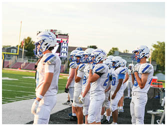 A group of people wearing white football uniforms and helmets stand in a line on the sidelines of a football field during a game or practice. Description generated by AI