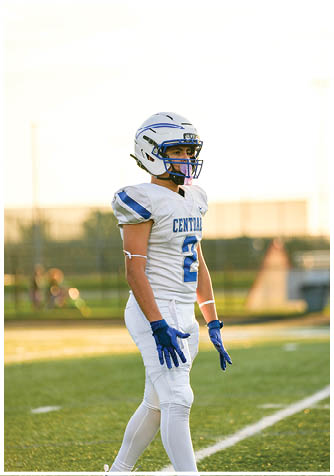 A person wearing a white and blue football uniform and helmet stands on a field with a focused expression during daylight. Description generated by AI