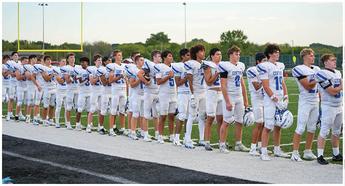 A group of people wearing athletic shoes are standing on a light-colored surface next to a dark track or field area with a blue and black backpack placed on the ground. Description generated by AI
