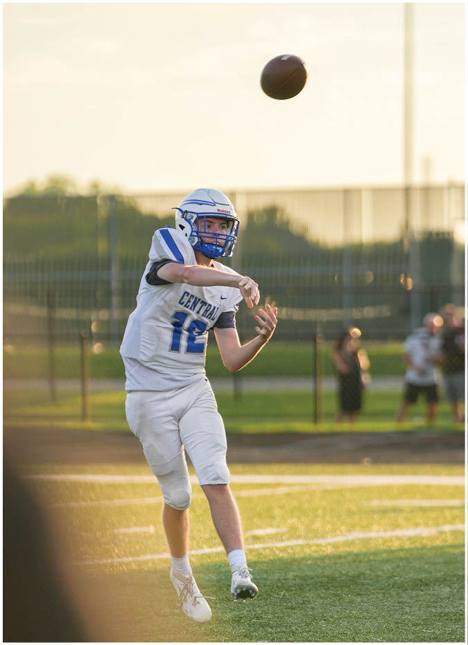 A person in a white and blue football uniform is throwing a football on a grassy field during sunset with other players and a fence in the background. Description generated by AI