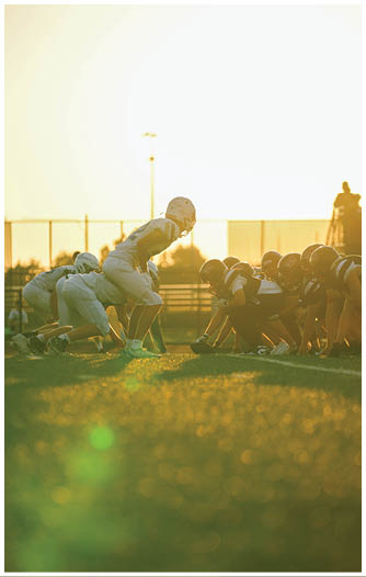 Two opposing American football teams in helmets and uniforms are lined up facing each other on a grassy field during sunset. Description generated by AI