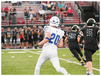 A football player wearing a white helmet with a blue logo is on the field with blurred spectators sitting in bleachers in the background. Description generated by AI