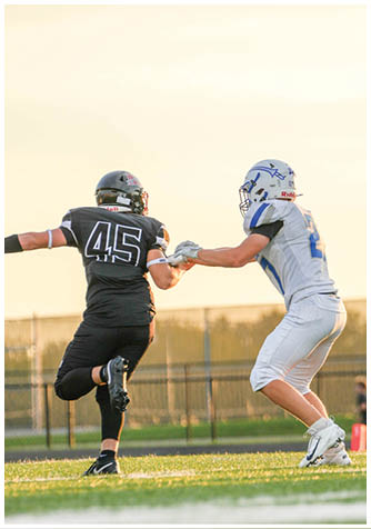 Two people in football uniforms are engaged in a play on a grassy field during sunset, with one person in black running while the other in white reaches out to block or tackle. Description generated by AI