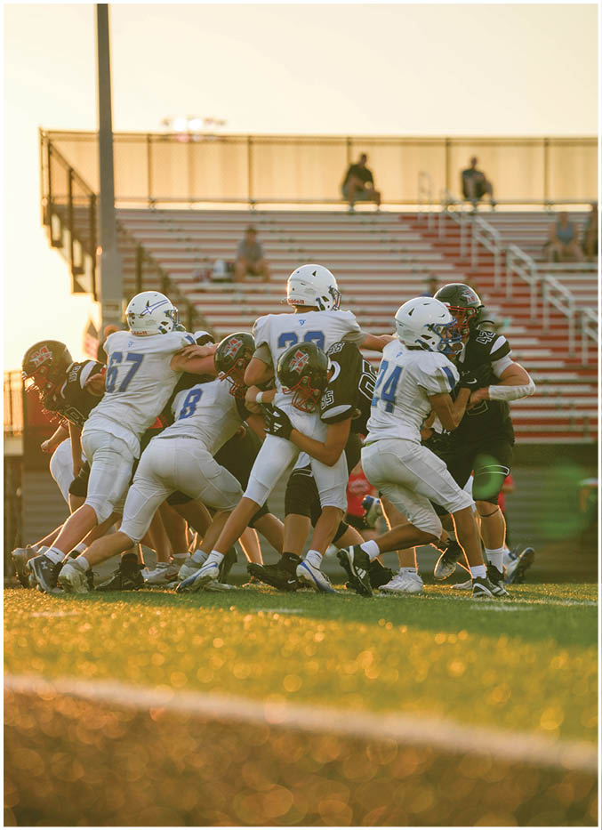 Several people wearing football helmets and uniforms are engaged in a close physical play on a football field with empty bleachers in the background. Description generated by AI