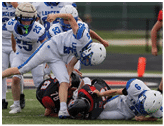 Two people in football uniforms are on the ground during a game, with one person in a white and blue uniform tackling another in a black and red uniform. Description generated by AI