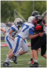 Two people in football uniforms are engaged in a tackle on a grassy field with other players visible in the background. Description generated by AI