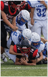 A group of football players in white and black uniforms are tackling a player in a black and red uniform who is holding the football on a grassy field. Description generated by AI