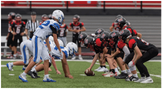 People in athletic shoes are positioned on a grassy field, appearing ready to start a football play with one person holding a football. Description generated by AI
