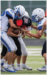 Three people in football uniforms are engaged in a tackle on a grassy field with one person holding the ball tightly while two others in white helmets and blue and white uniforms attempt to stop them. Description generated by AI