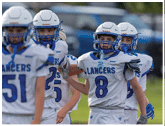 Children wearing white and blue football uniforms with helmets are standing together on a field, with one child in the center raising an arm. Description generated by AI