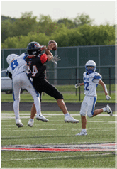 Two people in football uniforms are jumping and reaching for a football while another person in a football uniform watches nearby on a field. Description generated by AI