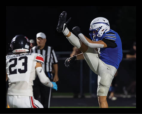 A person in a blue football uniform is kicking a ball while another person in a white football uniform with the number 23 watches during a nighttime game. Description generated by AI