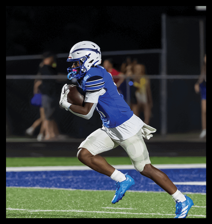 A person wearing a blue football jersey and white helmet is running on a field while holding a football. Description generated by AI