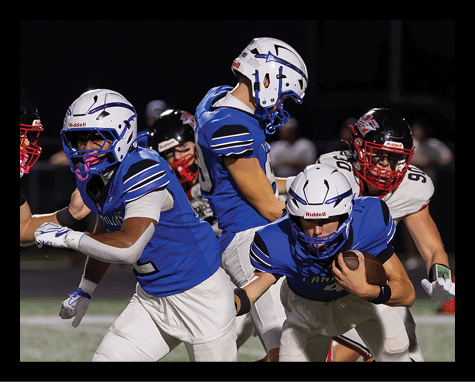 Several people wearing blue football uniforms and white helmets are gathered closely together on a field at night. Description generated by AI