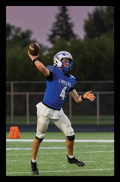 A person wearing a blue football jersey with the number 4 and a helmet is preparing to throw a football on a field with a fence and trees in the background. Description generated by AI