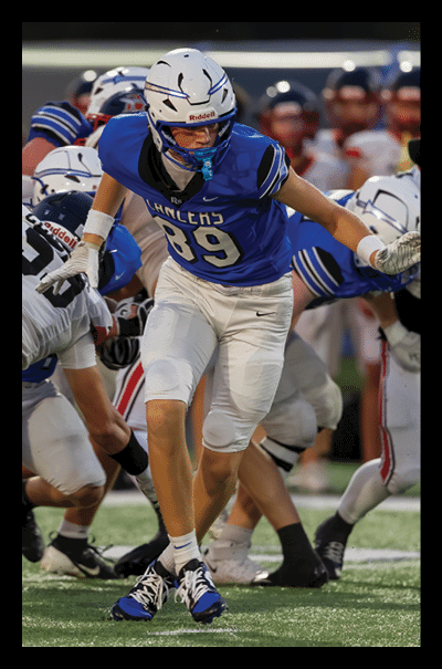 A person wearing a blue football jersey with the number 89 is running on a football field while other players in white and black uniforms are around them. Description generated by AI