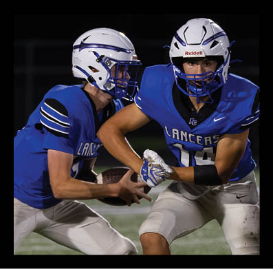 Two people in blue football uniforms and white helmets are engaged in a play on a football field at night. Description generated by AI